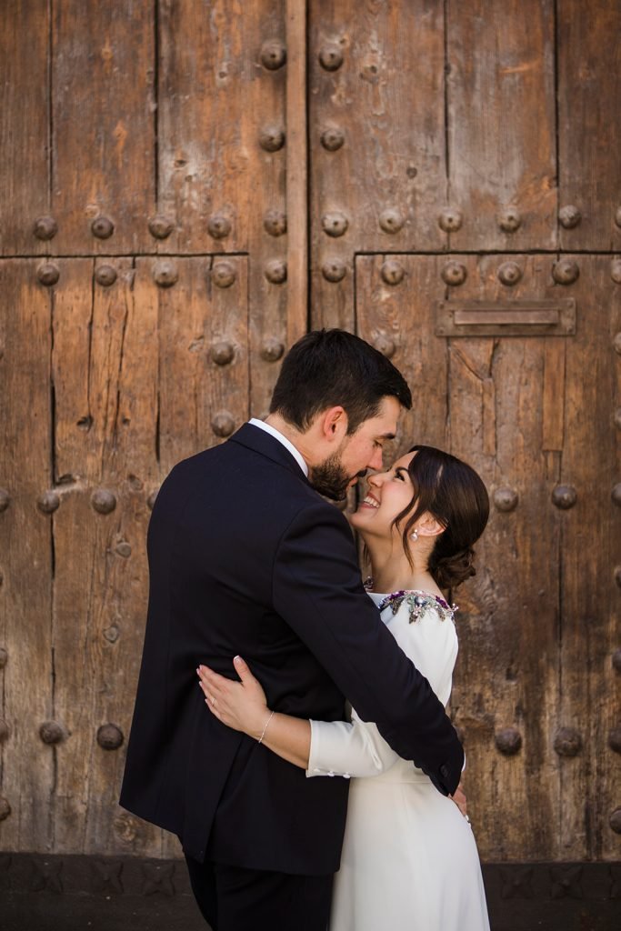 Romantic wedding couple portrait kissing in front of large historic wooden doors in Madrid. Editorial wedding photographer Madrid, Flor de Sal Photo.